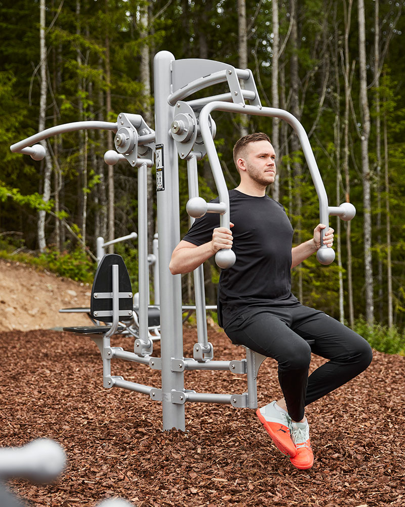 A man exercises at an outdoor gym station, he is surrounded by trees and nature.